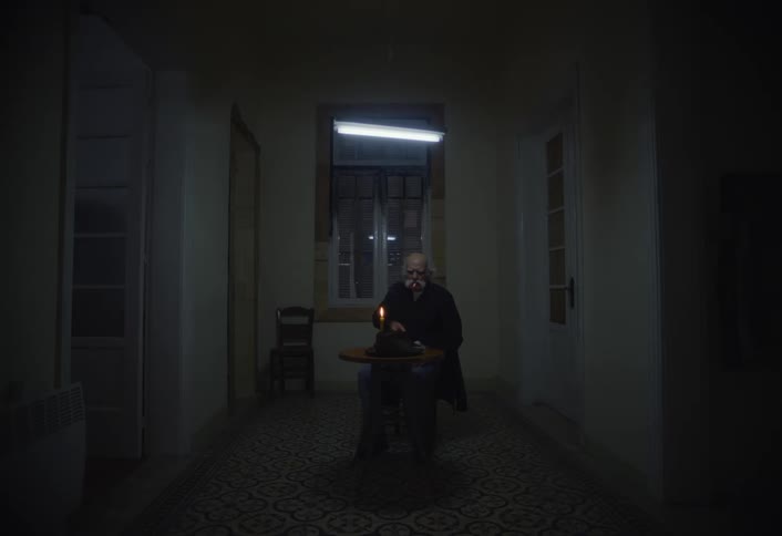 Elderly man sits at a small table in a nearly empty room at night and smokes