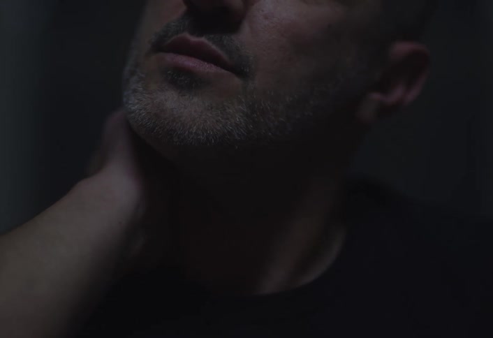 A man with short hair and a beard resting his hand on his neck and then his head with his eyes closed