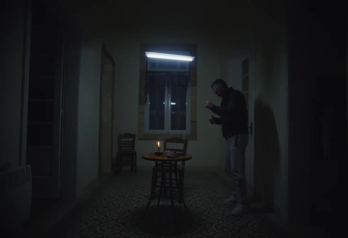 In a dimly lit room with patterned flooring a man jumps and shakes near a small table with a lit candle