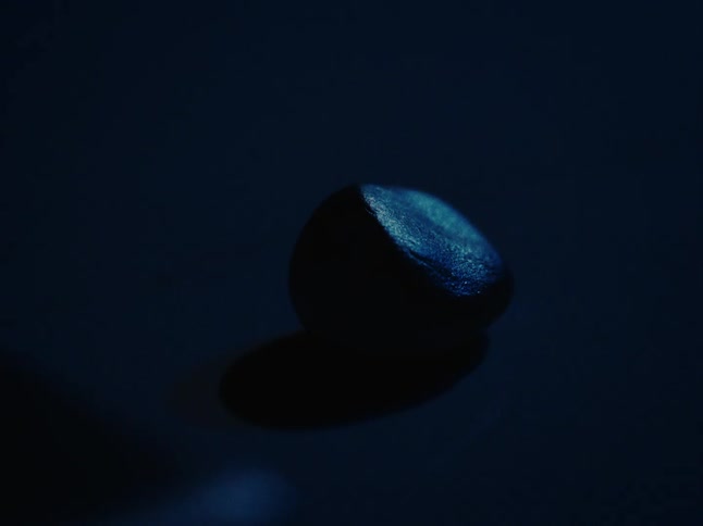 Fingers touch a small smooth darkcolored stone resting on a surface illuminated by blue light with a defined shadow cast below