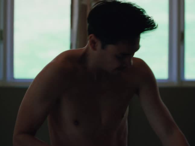 A shirtless man dances energetically alone in a dimly lit room near a window