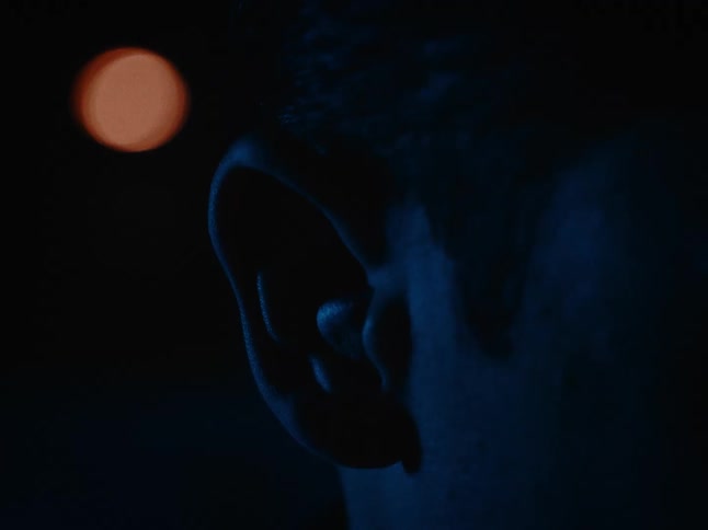 A closeup dimly lit shot shows the side of a person placing an earbud in their ear with a blurred light in the background