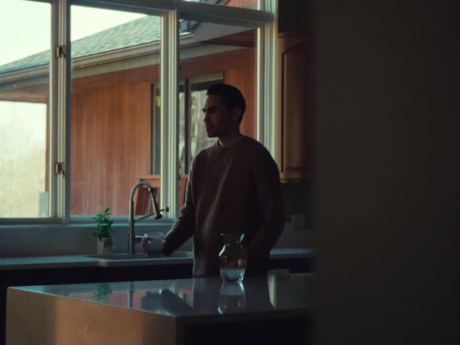 A person stands at a kitchen sink serving a glass of water near a large window with a view of a house exterior