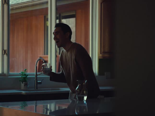 A lone man stands still in a kitchen holding a glass of water lets out a brief laugh then straightens up to take a drink in an odd manner