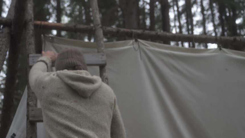 A person wearing a beanie and sweater adjusts a canvas shelter supported by wooden poles in the forest
