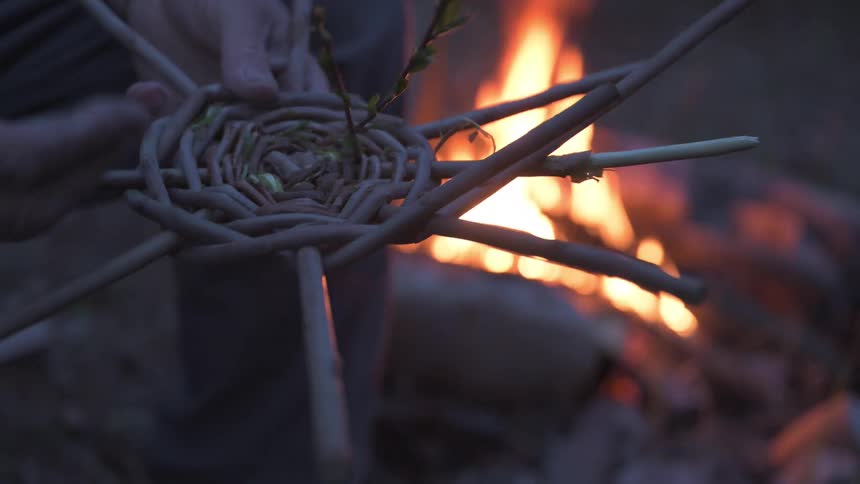 A closeup of a persons hand working on a handcrafted wicker basket near a campfire at dusk