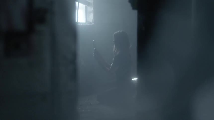 A woman is kneeling on the ground in a room and raising a mirror up to her face