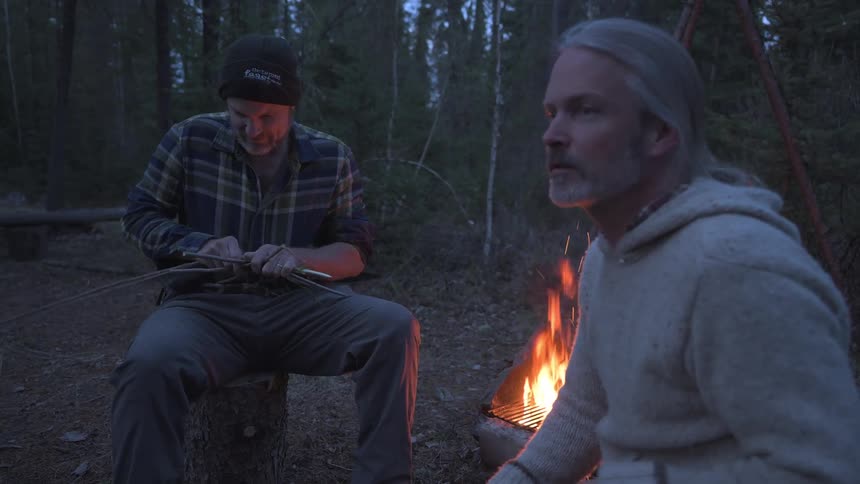 Older male friends talk while sitting in the woods near a campfire