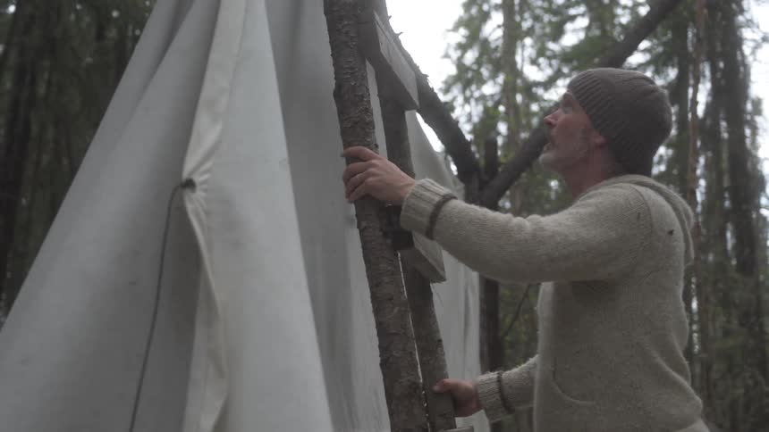 A person wearing a beanie and sweater adjusts a canvas shelter supported by wooden poles in a forest