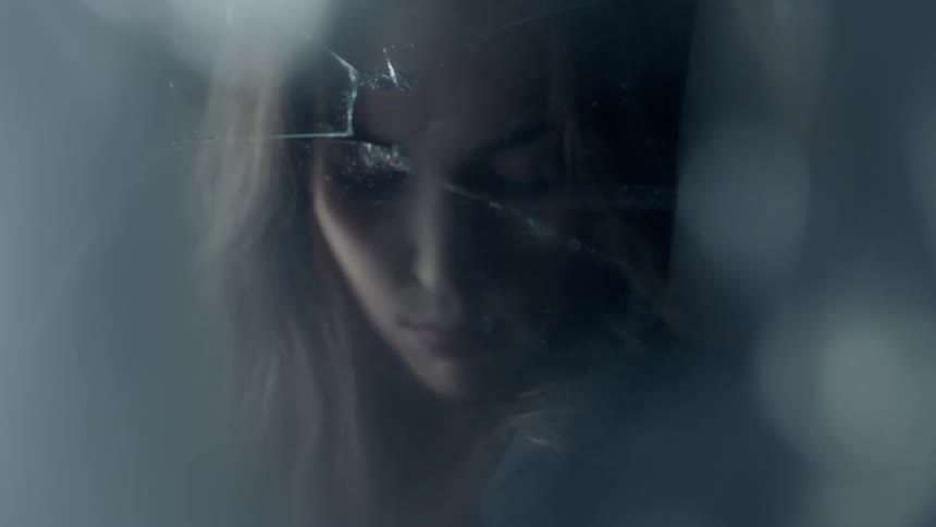 A woman is standing before a broken mirror with her eyes cast down