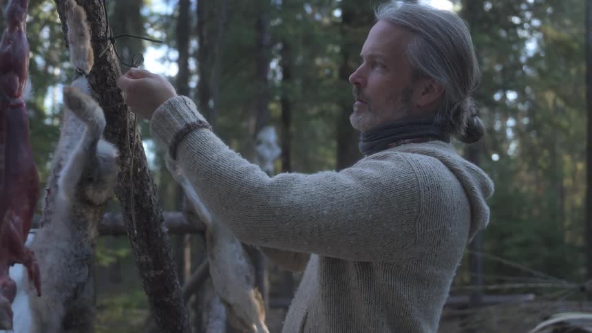A man in a wool sweater examines several skinned rabbits hanging from a branch in a wooded area