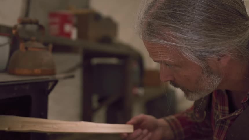 A person places wood into a black stove with a kettle and a white container on top inside a tent