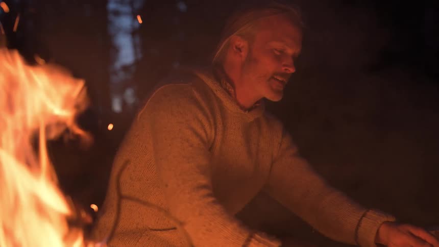 The campfire rages as the man talks