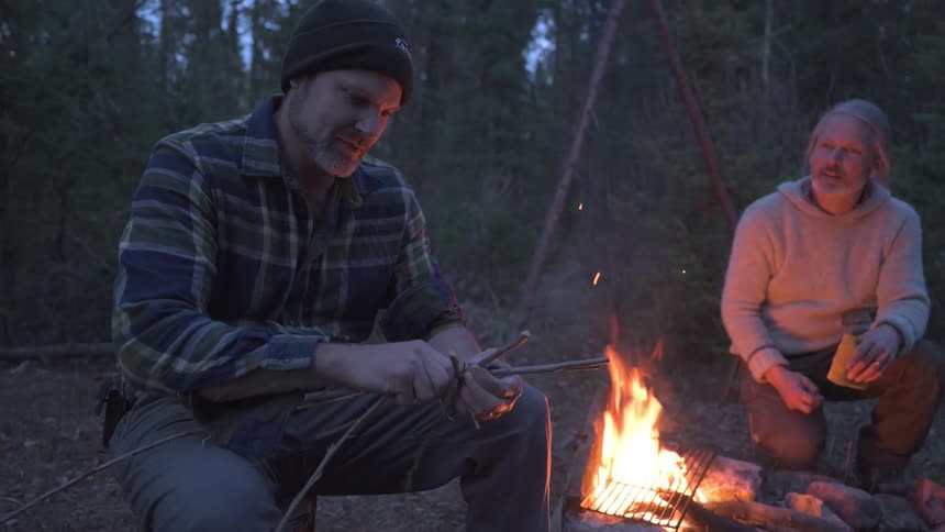 A man is tying sticks and talking with a friend at the campfire