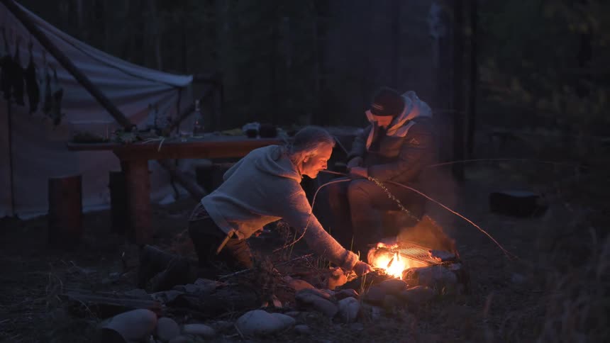 A hunter tends a small fire in a dark outdoor setting near a tent with a person weaving a basket nearby