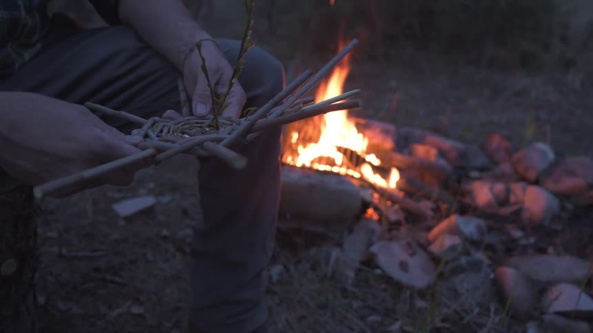 A man is holding an object made of sticks by the campfire