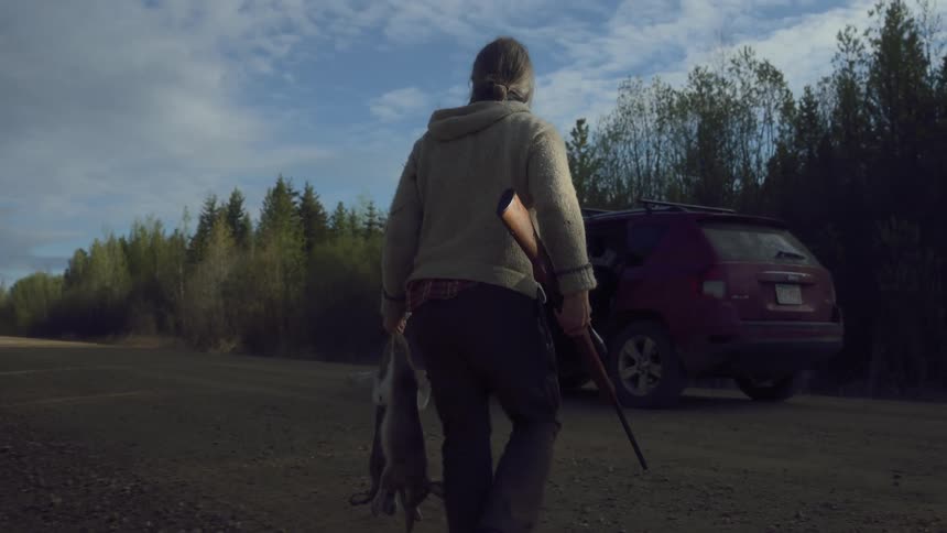 A hunter carrying a rifle and two dead rabbits emerges from the woods toward a car parked on a dirt road in the morning