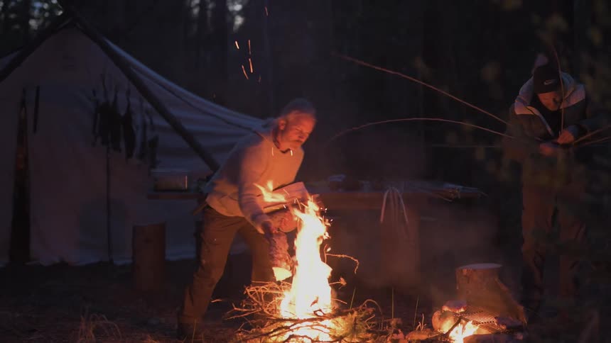 Two people tend a campfire at night in a forest one adding logs and the other threading a baket