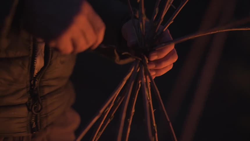 A person holding a bundle of branches the person looks as if they are tying the branches with a thinner smaller plyable branch the shot is taken at night and has a warm orange glow as if the light of the shot is coming from a campfire