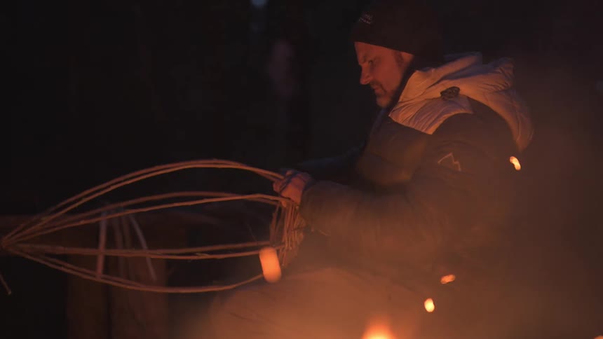 Two men are in a forest at night one feeding a campfire while the other works on handcrafting a wicker basket