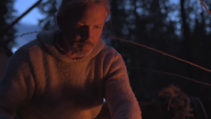 Two men work on their tasks while sitting near a campfire in the evening