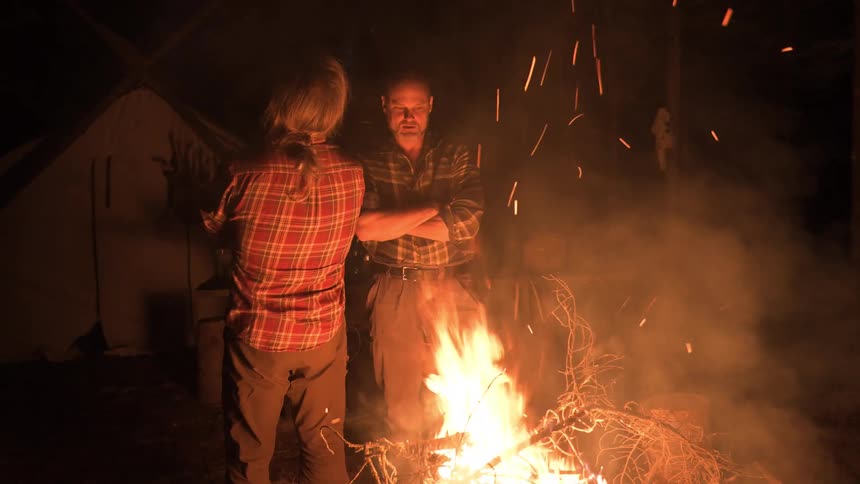 Two men stand around a brightly burning campfire at night with sparks and smoke illuminating the darkness