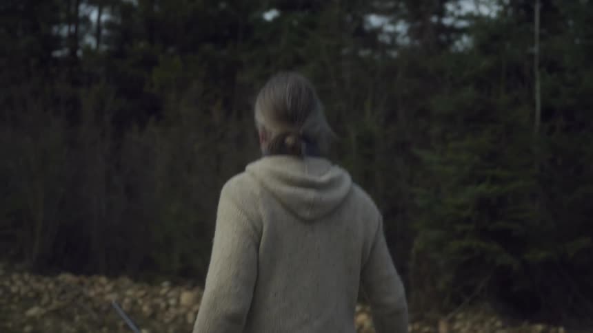 A man with long gray hair wearing a gray hoodie and dark pants walks away from the viewer carrying a rifle in a dark wooded or brush filled environment