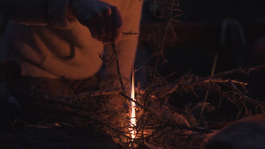A closeup of a mans hand using small twigs as tinder to ignite a campfire at night