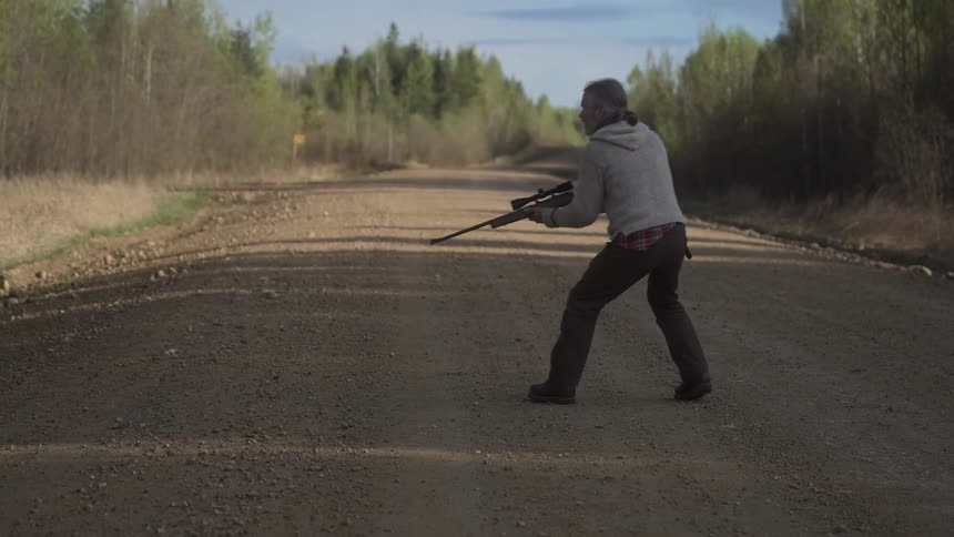 A hunter kneels on a dirt road aiming a rifle with a scope towards a target
