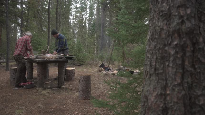 Two individuals process game at a rustic table in a forest clearing near a fire pit