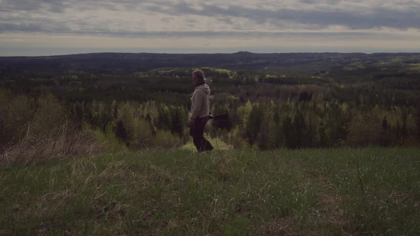 A hunder walks across a grassy field carrying a rifle against a backdrop of rolling hills and a dense forest