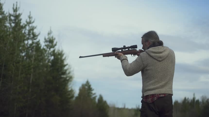 A man with long gray hair wearing a lightcolored sweater and dark pants aims a rifle with a scope in a wooded area