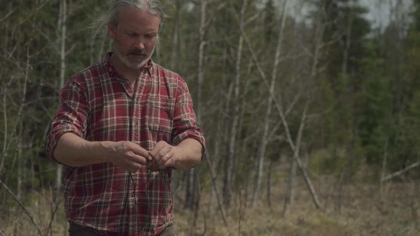A man in a plaid shirt manipulates a thin wire in a forest with sparse trees