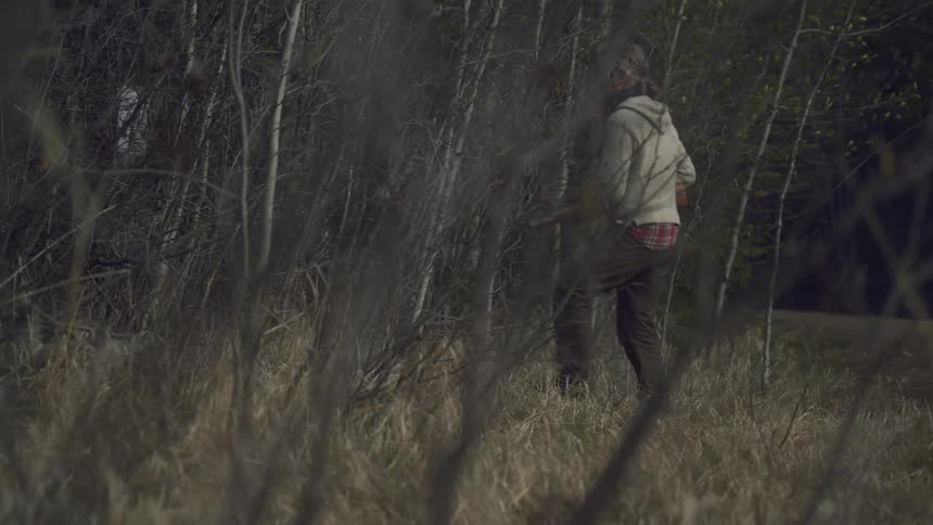 A hunter with long gray hair wearing a light colored sweater and dark pants walks along a dimly lit dirt road at dusk carrying a rifle with a dark forest in the background