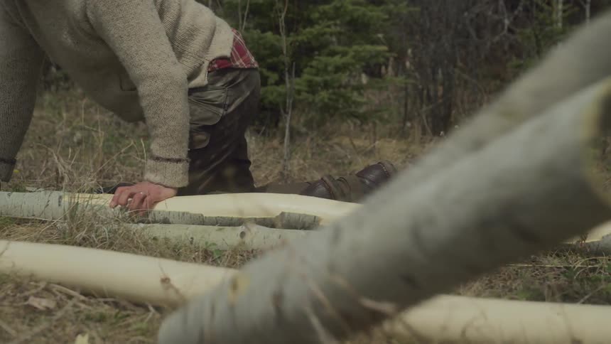 Two men sit near a wooded area stripping bark off of tree trunks with an ax