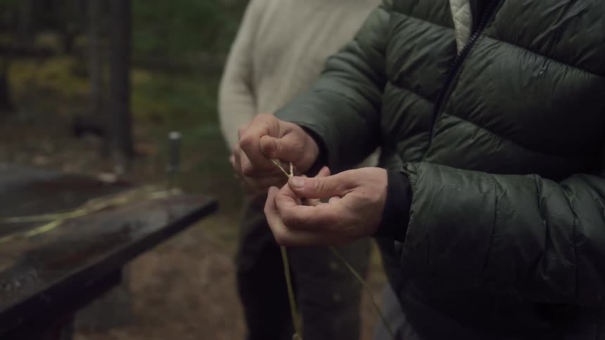 A man observes another crafting a length of twine in the forest engaging in handcraft and woodcraft