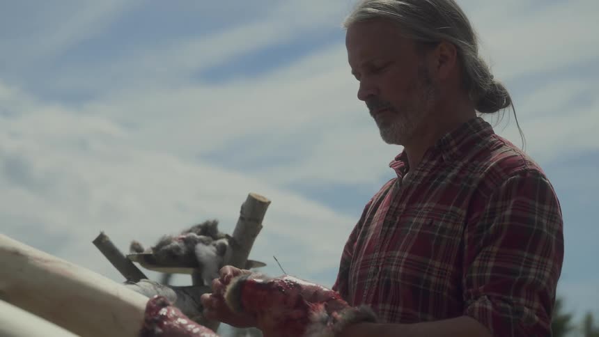 A man in a plaid shirt stands outdoors under a partly cloudy sky working with animal hides stretched on a wooden frame