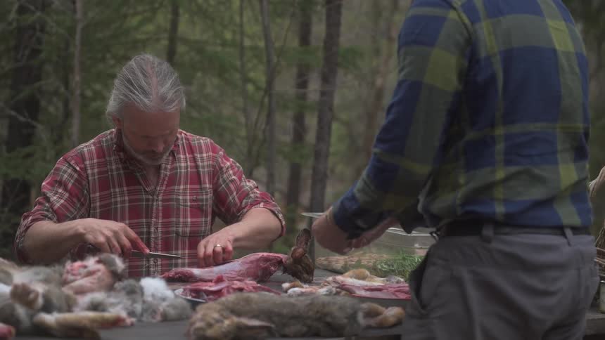 A couple of hunters process a rabbit carcass peeling fur with knives at a table in a rustic forest setting