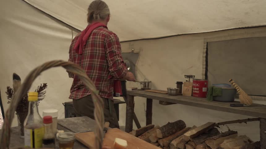 A man is inside a large tent cooking and placing a pot on a wooden stove