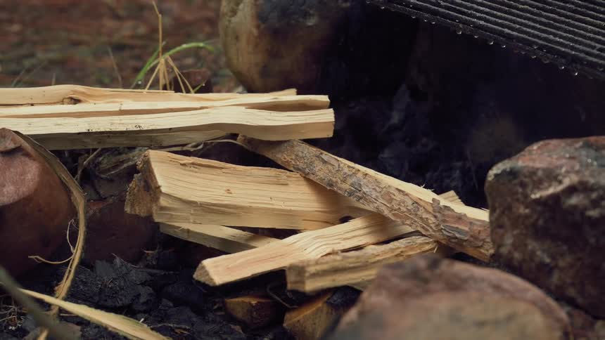 A mans hand carefully arranges pieces of firewood in a fire pit surrounded by rocks and charcoal