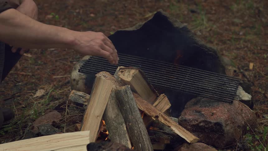 Hands place a piece of wood onto a small fire burning under a metal grate surrounded by rocks