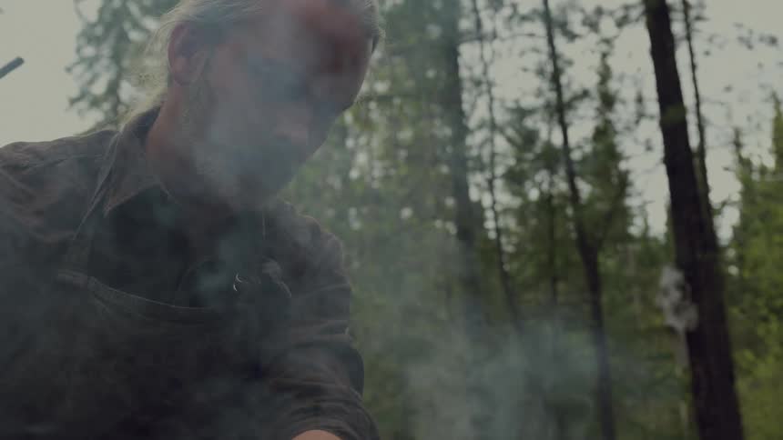 Smoke rises as a man puts branches into the campfire