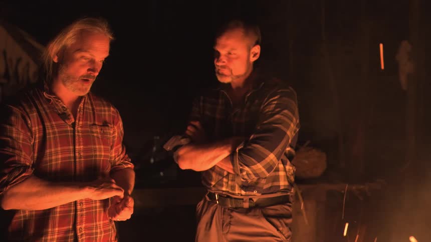 Two men stand side by side in a dimly lit rustic setting possibly outdoors with their arms crossed and serious expressions