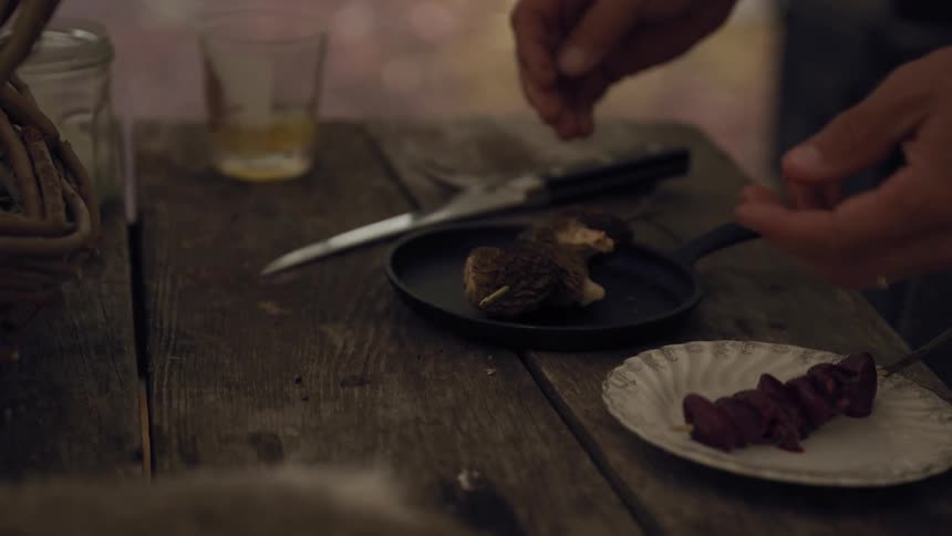 A man assembles meat brochettes on skewers over a small iron pan placed on a rustic outdoor table