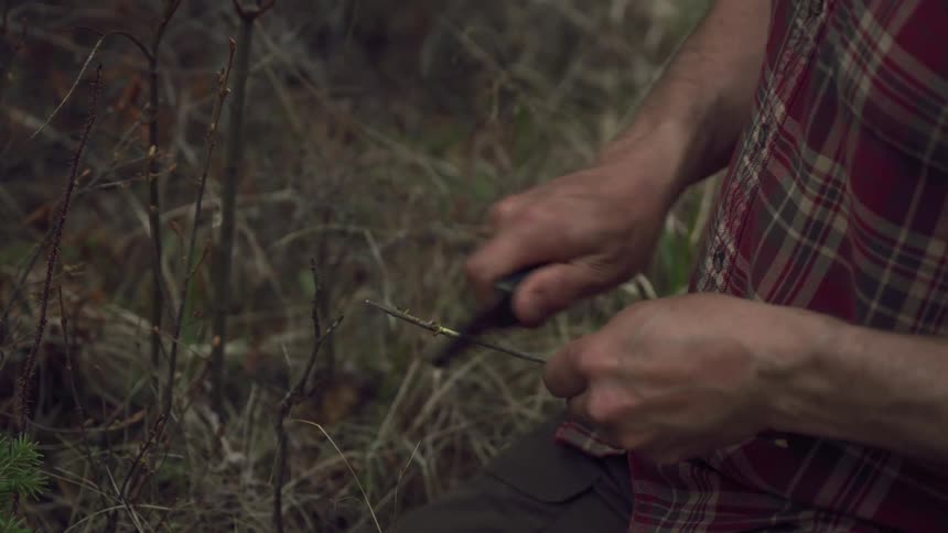 Man uses a knife to shave a stick while outdoors during the day