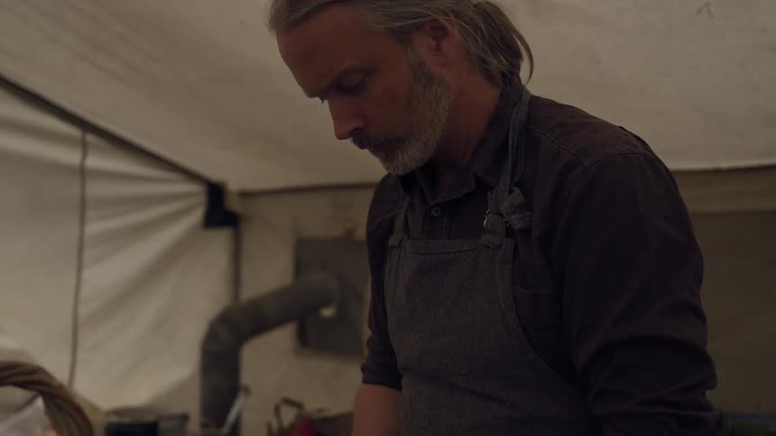 Older man prepares to cook the rabbit he caught while standing in a large tent near a standalone wood stove