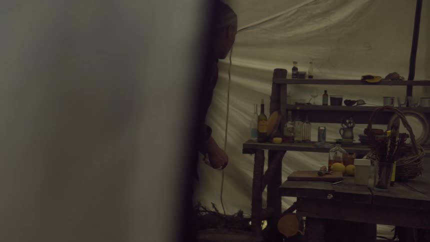 A man in a plaid shirt retrieves items from a rustic wooden shelf filled with various objects inside a tent