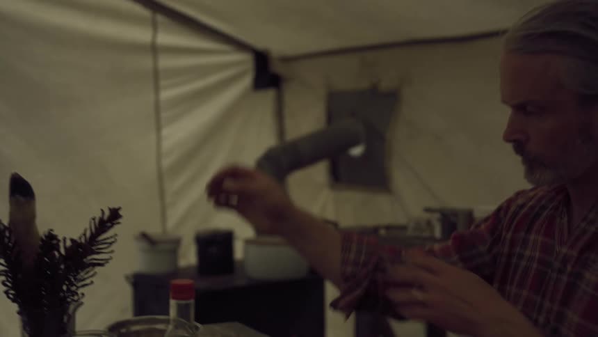 A camper stirs a pot and opens a jar as he cooks in his tent
