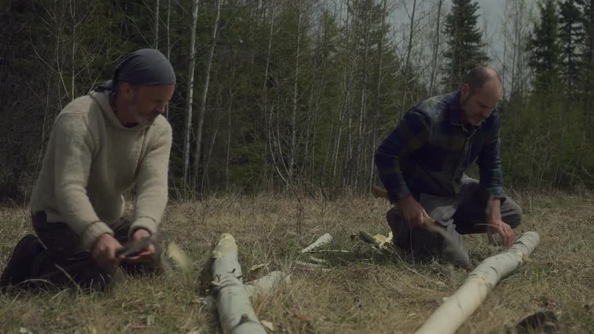 Two individuals kneel in a wooded area using axes to cut and shape large logs