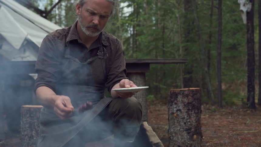 A man kneels in front of a grill over a campfire placing a meat brochette surrounded by a forest
